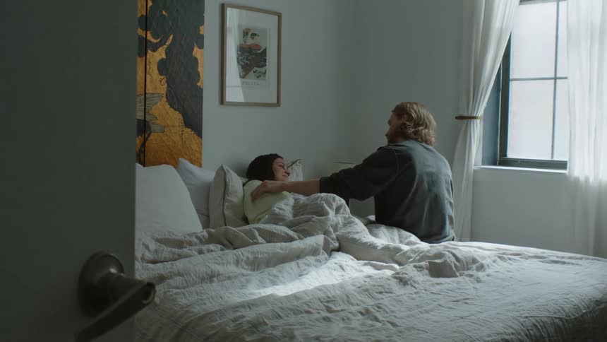 A man sits on the side of a bed comforting a female laying in bed wrapped in the sheets