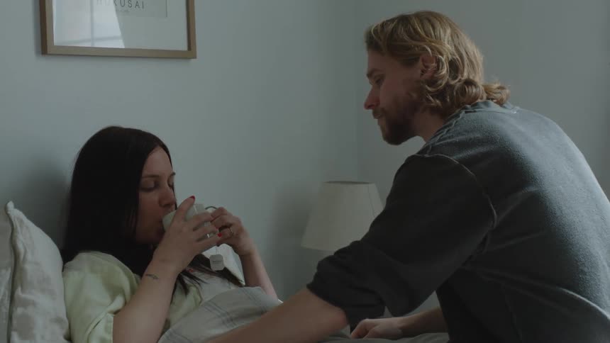 Man gives cup of tea to woman lying ill in bed before he sits and strokes her arms