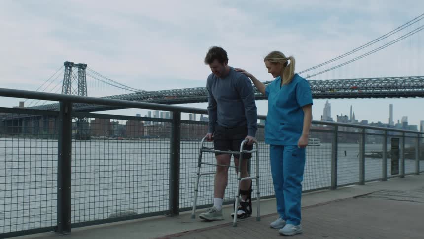 A nurse walks next to a man with his foot in a brace as he uses a walker and she rests her hand on his shoulder