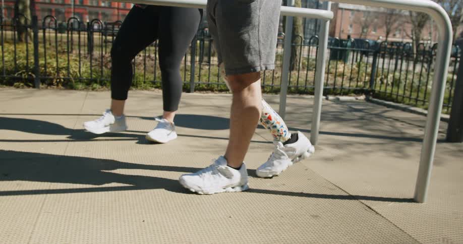 Therapist helps man with a colorful prosthetic leg to walk in a park near handrails
