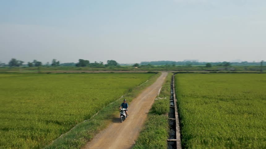Man rides motorcycle along dirt road between crop fields with mountains in the background