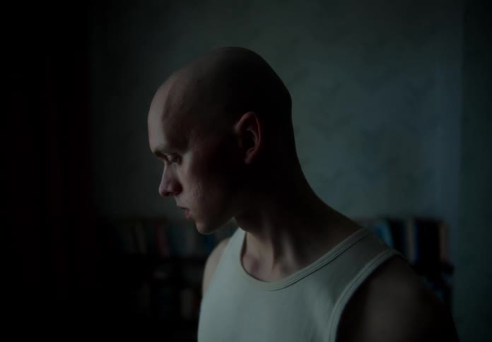 Bald man with white undershirt stands in dark room