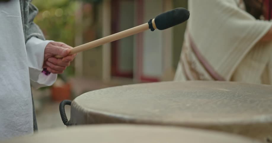 A persons hands are hitting a large barrel drum with drumsticks while a woman wearing a shawl is off in the corner
