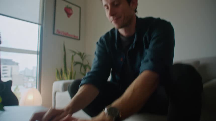 A close up of a man sitting on the edge of his couch typing on the laptop in front of him