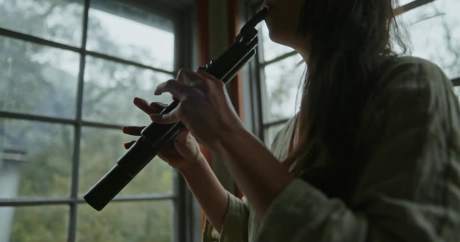 A woman stands indoors as she plays a wooden wind instrument