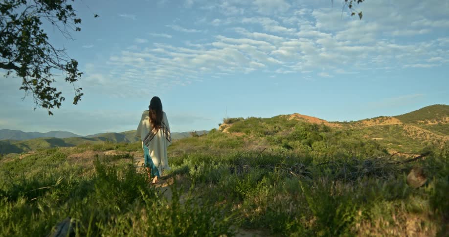 Woman with brown hair and white shirt walks along dirt track in the field