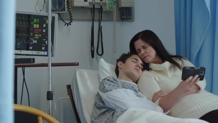 A mother sits on the edge of a hospital bed next to her son holding a cell phone out while watching it while the song sleeps on her shoulder