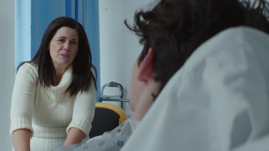 Woman with dark hair and white sweater talks with patient lying in bed at the hospital