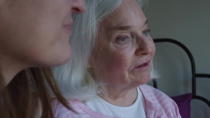 Elderly woman and her daughter sit on bed talking together