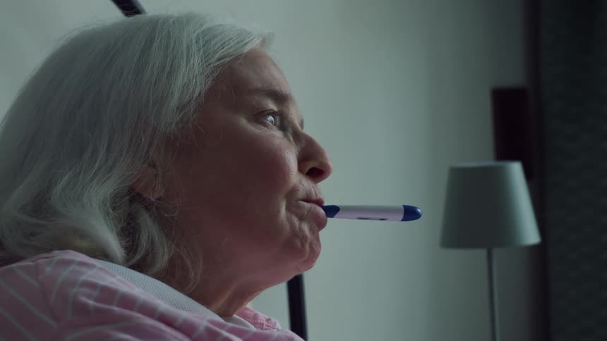 A close up of a person putting a thermometer in and elderly womans mouth as she nods her head as it is taken out