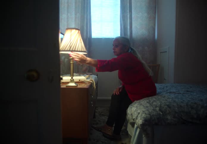 Elderly woman with ponytail red shirt and black pants sits on bed turning off bedside lamp