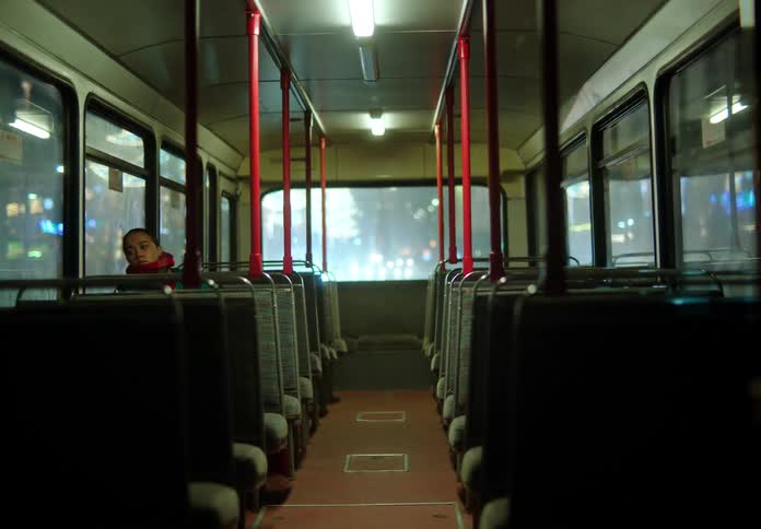 A woman bundled up in winter clothes is the only passenger sitting on a bus as it travels on a city street