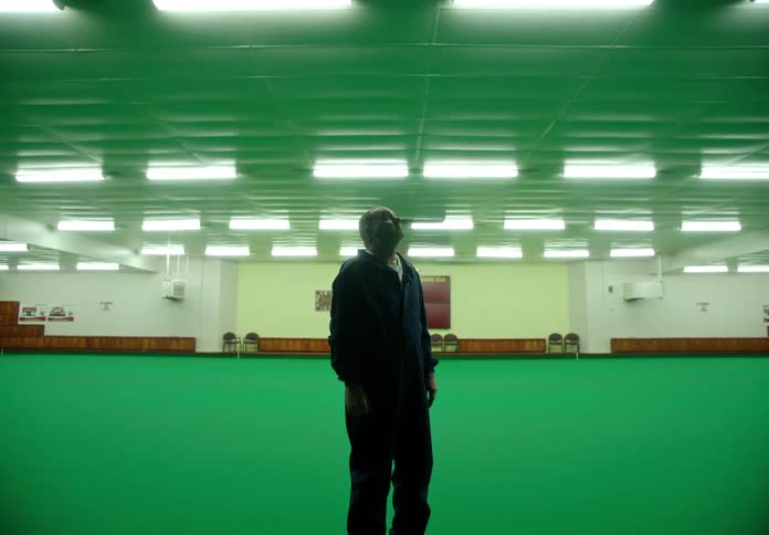 A man in a blue jumpsuit stands in the middle of a large greencolored room looks up hopelessly and slowly puts his head in his hands