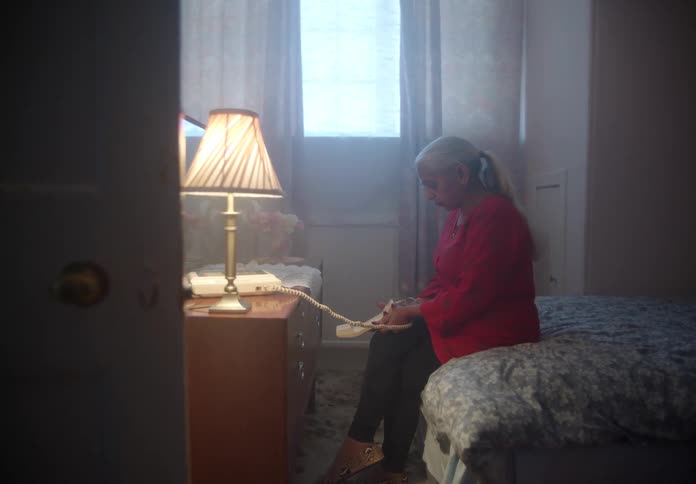 An elderly woman sits alone in a bedroom while holding a phone receiver in her hand