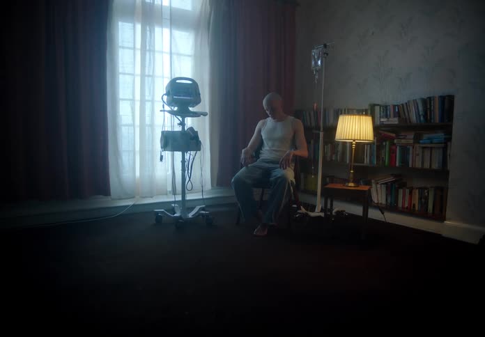 Young bald man sitting on a dimly lit room at home surrounded by medical equipment