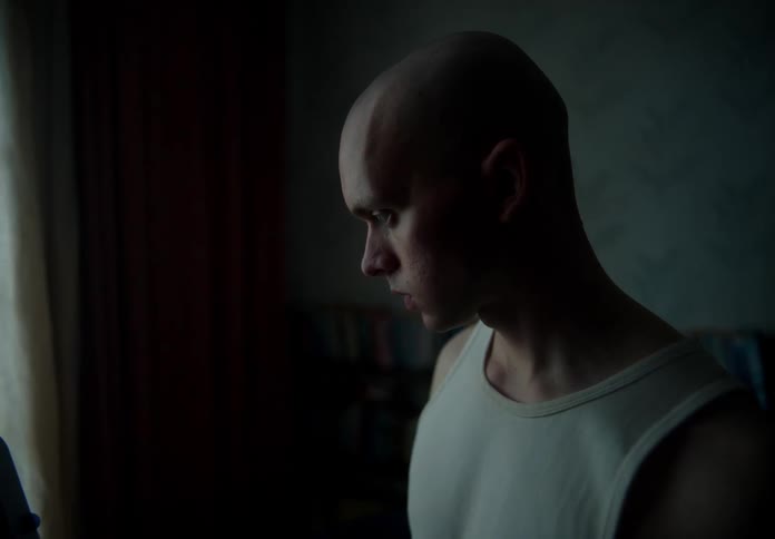 Bald man with white undershirt looks down in dim lit room