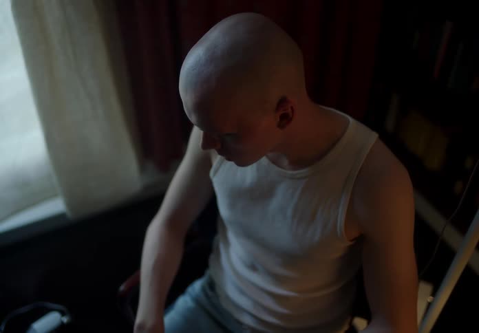 Bald man with white undershirt and jeans sits on chair in room