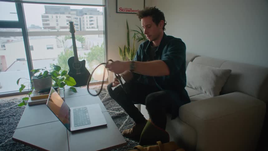 Man walks through kitchen and sits on couch in living room before opening laptop and looking at camera
