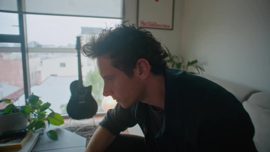 A man is sitting on his living room couch as he take his camera out of a bag and looks at the laptop computer on the table in front of him