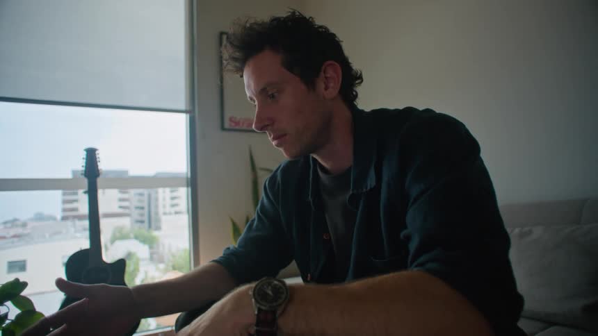 Man in black shirt and pants sits on gray couch and makes a video call on laptop while holding camera in the living room
