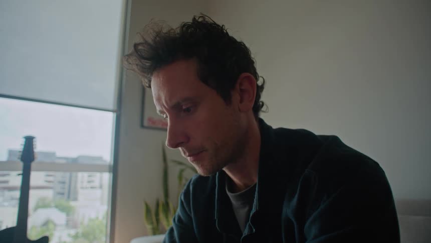 Man in black shirt and pants sits on gray couch types on laptop in the living room