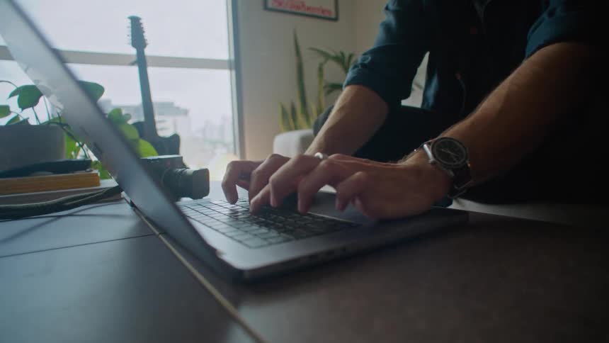 A man is sitting at a table typing on a laptop computer and he stops to think