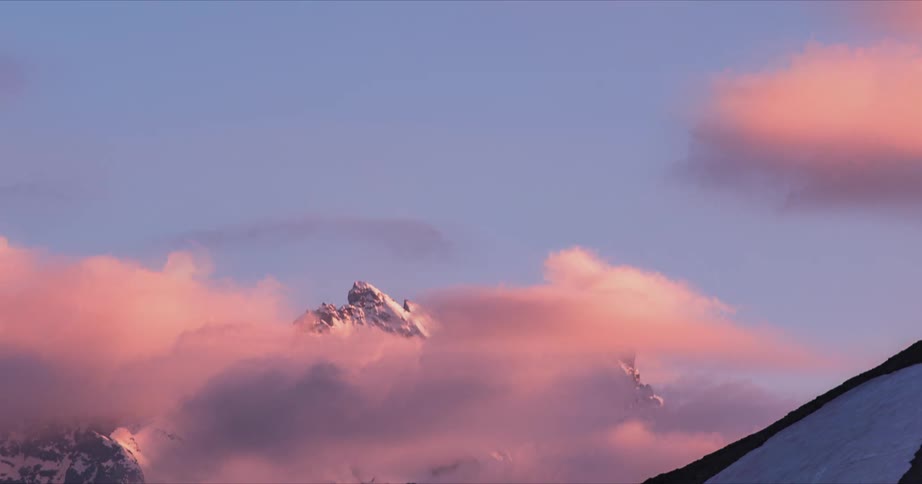 A time lapse of clouds moving past snow covered mountain peaks as dusk sets in
