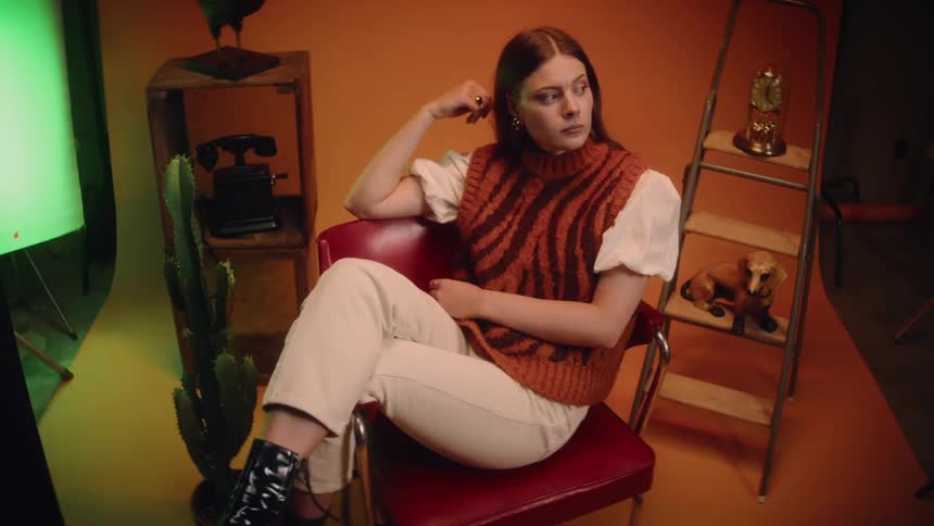 A woman sitting in a chair with her legs over the arms of her chair with decorations behind her on an orange backdrop