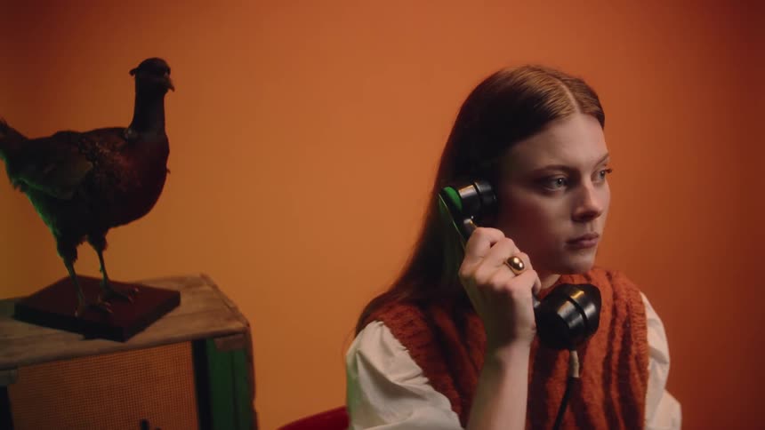 A woman is sitting in a chair in a room with deep orange walls as she picks up a big old fashioned telephone receiver as she answers the phone and puts it back down