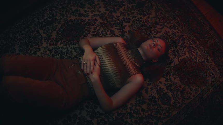 A woman laying on her back on the floor with her hands on her stomach as she looks up at the ceiling as shadows cast across her