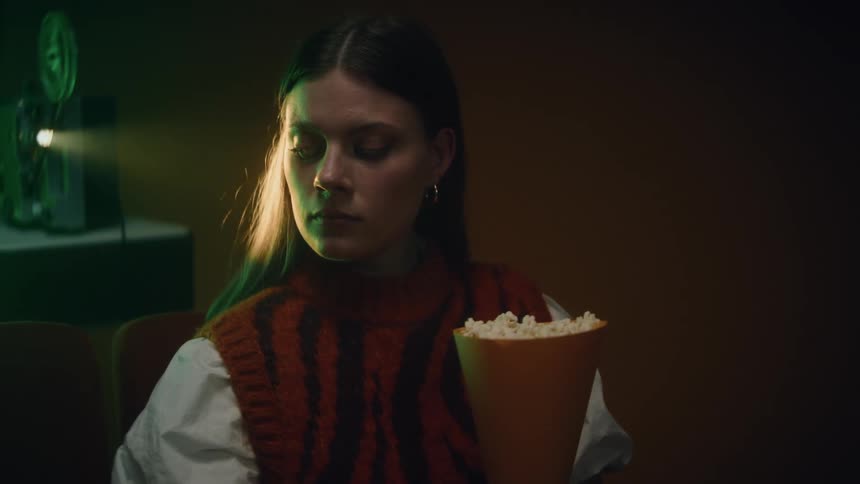 A woman holding popcorn sits in front of a movie projector while glancing around the darkened room