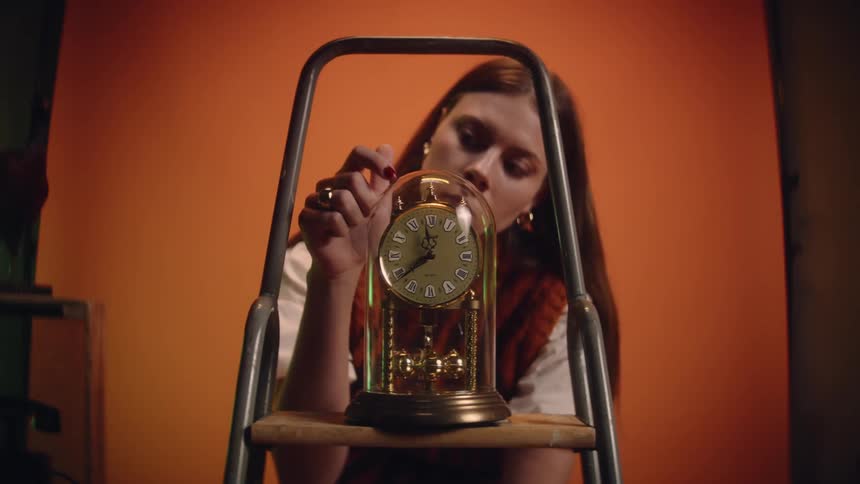 A woman sitting in front of an orange backdrop looking at an old clock in a glass on a wooden shelf