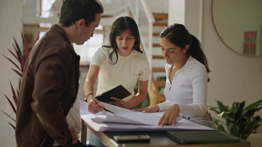 Three people are standing over a table in a room looking at plans as they talk and one woman is taking notes