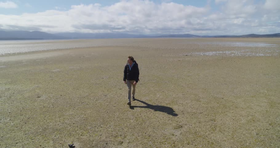 A man wearing a backpack and a jacket is walking across wide open land and there are mountains in the distance under a cloudy sky