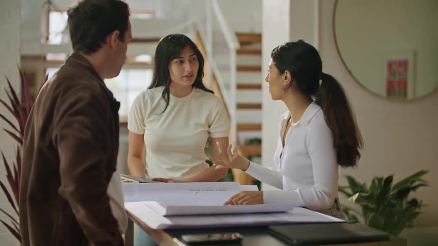 Three people are standing over a table in a room looking at plans as they talk and one woman is taking notes