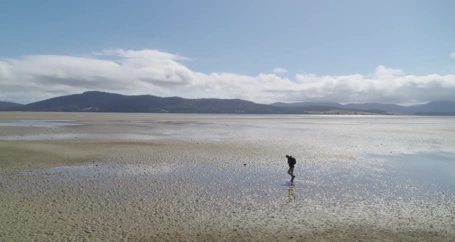 An aerial view of a man walking through muddy wet sand and a landscape view of the flat and mountain range in the distance