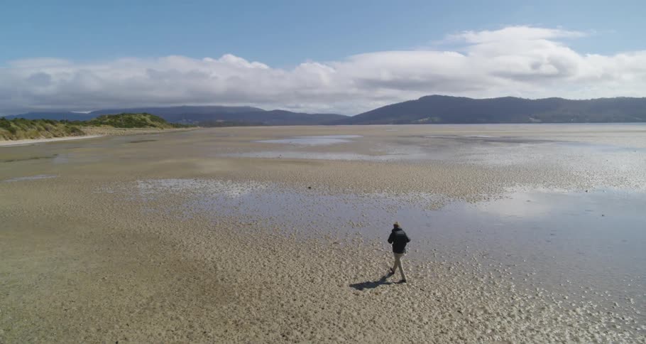 An aerial view of a man wearing a backpack walking across wet sand with mountains in the far distance
