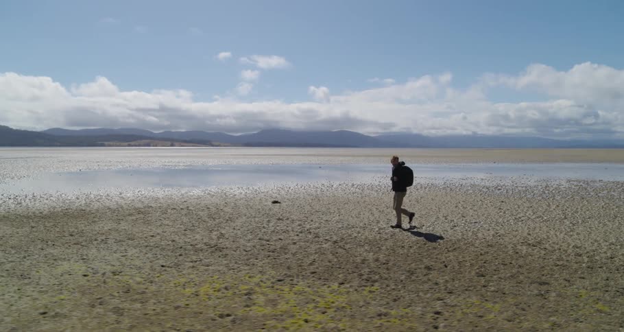 Man with backpack jacket and brown pants walks across dried lake