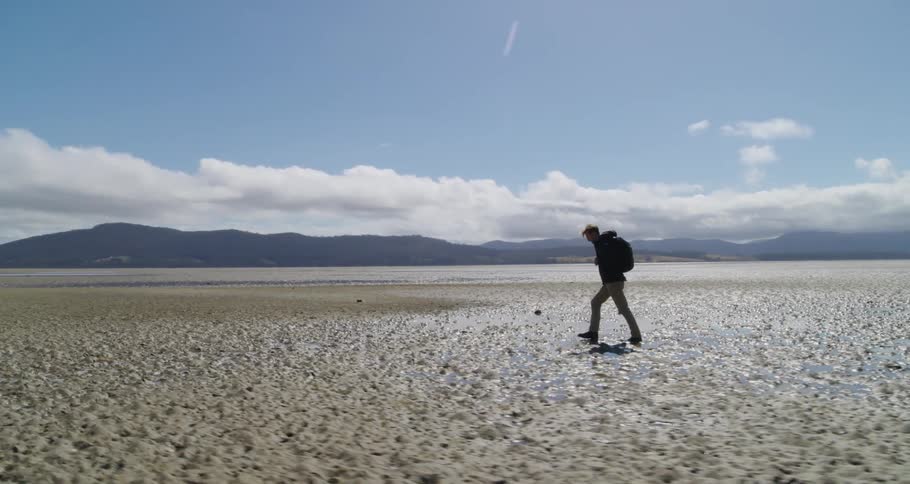 A man walking through a wet sandy flat land with mountain silhouettes in the distance