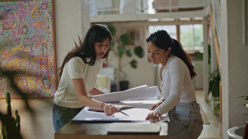 Two women are standing over a table looking at plans and pointing at notes