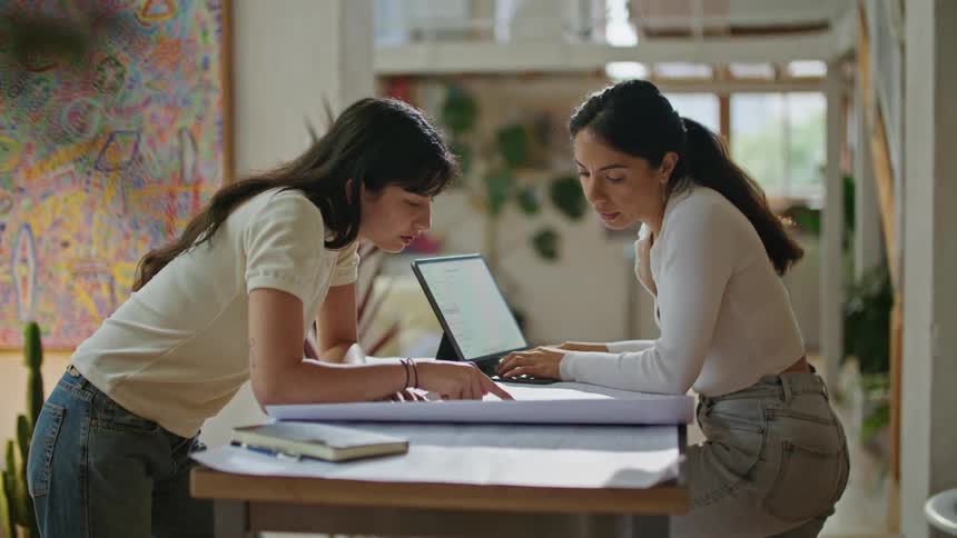 Two women review a large paper while conversing an typing on a laptop
