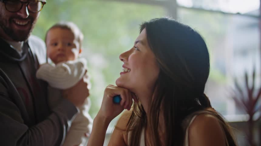 A man wearing glasses holding a baby while he talks to his wife sitting in front of them who is laughing and smiling