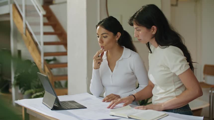 Two women go over blueprints while consulting using a laptop