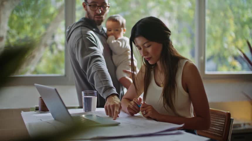 Father with glasses beard and gray hoodie stands next to mother working on laptop holding baby in his arms