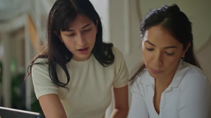 Two women are standing over a table with a computer as they look over plans and one woman is pointing as they talk