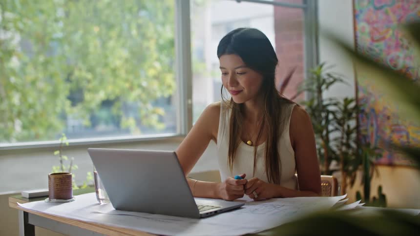 A woman is sitting at a desk in front of a laptop computer as she waves to the screen in a video conference