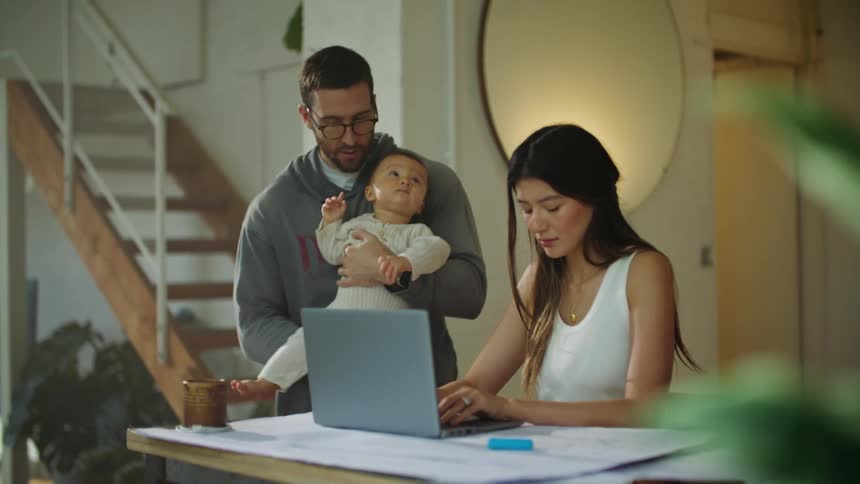 Mother with white shirt works on laptop while dad rocks baby in his arms