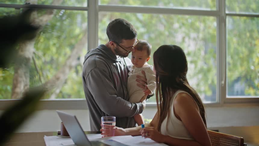 A woman sitting at her home desk looking at her laptop when her husband brings her a glass of water carrying their baby