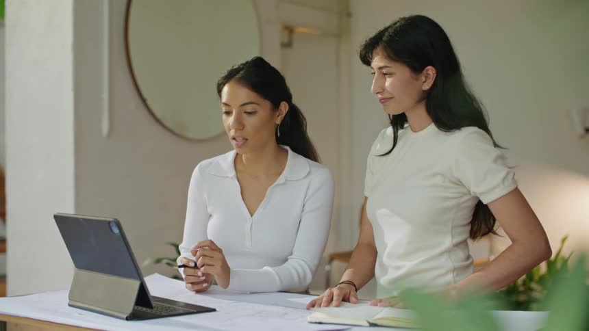 Two women participate in video meeting while standing at table consulting a book and taking notes