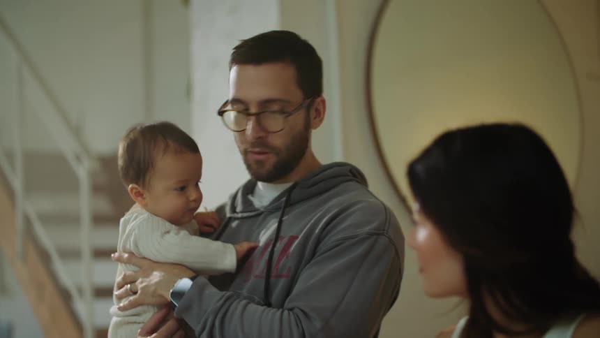 Father with beard and hoodie holds baby while mom using laptop
